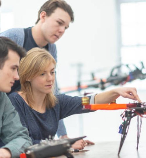 Four classmates are working at a table. They are looking at a completed drone standing on the table. There is another drone flying in the background.