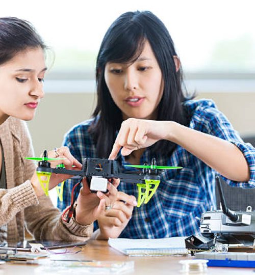 Pretty Asian teacher at STEM school helps Hispanic student with drone in engineering class. The teacher is pointing to something on the drone as the student looks on. The controller and other parts or tools are on the table. The teacher is wearing a blue plaid shirt and the student is wearing trendy layered clothing.